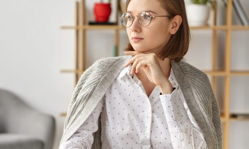 Portrait of thoughtful beautiful woman generates new ideas for future project, tries to gather with thoughts, keeps hand under chin, dressed elegantly, sits over home interior. Successful woman indoor